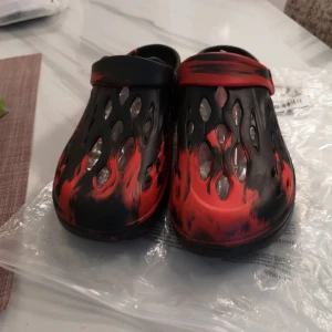 Dual-Tone Clog Sandaler - Säljer ett par sandaler i svart och rött med unikt, eldliknande mönster. Skorna har öppningar på ovansidan för extra ventilation och en justerbar rem bak. Tillverkade i syntetmaterial, perfekta för chill dagar eller när du vill sticka ut lite extra.