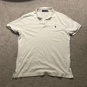 Vit pikétröja från Polo Ralph Lauren - Klassisk vit pikétröja från Polo Ralph Lauren med broderad marinblå logga på bröstet. Tröjan har korta ärmar, krage och knäppning framtill. Tillverkad i mjuk bomull som känns skön mot huden. Perfekt för en clean och stilren look.