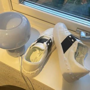 Valentino Garavani vita sneakers - Snygga vita sneakers från Valentino Garavani med svart band och nitar på hälen. Skorna har rund tå, platt sula och klassisk snörning. Tillverkade i skinn för en lyxig känsla och stilren look. Perfekta för dig som vill sticka ut med exklusiva detaljer.