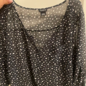Svart blus med hjärtan från Torrid - Säljer en svart blus från Torrid med vita hjärtan över hela tyget. Blusen är tunn och luftig med v-ringning, knappar framtill och bröstficka. Perfekt för dig som gillar söta detaljer och vill ha en lätt och flowy topp.