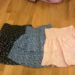 3-pack blommiga volangkjolar - Säljer tre söta korta kjolar med volanger och smock i midjan. En är ljusrosa med vitt blommönster, en blå med små vita blommor och en svart med vitt blommönster. Perfekta för sommarens alla dagar och superbekväma med stretchig midja.