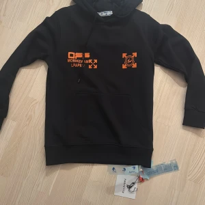 Svart hoodie Off White x Moncler  - Svart hoodie med orange och gula tryck från Off Moncler x PlastiU. Stor logga och grafiska detaljer på ryggen, samt mindre tryck framtill. Huva med snörning och klassisk känguruficka. Perfekt för dig som gillar streetwear vibes.