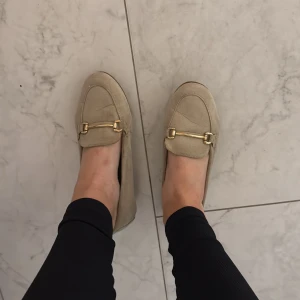 Beige mockaloafers med gulddetalj - Snygga loafers i beige mocka med guldfärgad metallspänne framtill. Skorna har platt sula och rund tå, vilket ger en klassisk och stilren look.  Lite slitna därav schysst pris, inget fel i helhet men man ser ”veck”, och är lite mörkare där hälen har varit på insidan. Köparen står för frakt & paket skickas per post :)