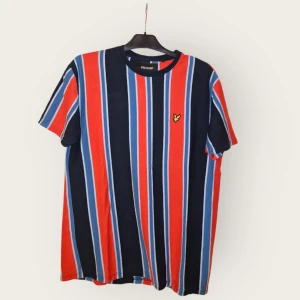 Randig t-shirt från Lyle & Scott - Snygg boxy fit t-shirt från Lyle & Scott med breda vertikala ränder i rött, blått och svart. Klassisk rund halsringning och korta ärmar. Ikonisk gul logga broderad på bröstet. Perfekt för dig som gillar färg och streetstyle.
