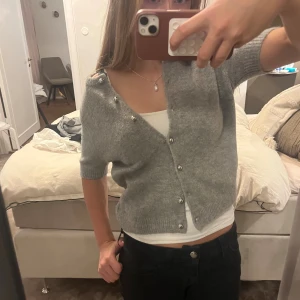 Grå kofta med knappar från zara - Mysig grå kofta från Zara i ullmix med korta ärmar och rund hals. Koftan har ribbade muddar och knäpps framtill med silvriga knappar. Perfekt för lager-på-lager och enkel att styla till olika looks.