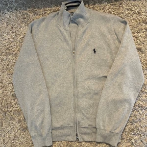 Grå kofta från Polo Ralph Lauren fullzip herr M  - Klassisk grå kofta från Polo Ralph Lauren med hel dragkedja och broderad logga på bröstet. Tröjan har ribbade muddar och är tillverkad i mjuk bomull. Storlek M i mycket bra skick. Postas antigen samma dag som köp eller dagen därpå🙌💯