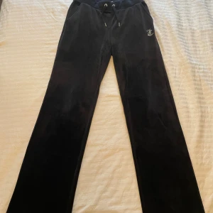 Svarta velourbyxor Juicy Couture S - Svarta mjukisbyxor från Juicy Couture i mjuk velour med snörning i midjan och fickor. På ena höften finns en glittrig JC-logga och på baksidan står det 'Juicy' i strass. Byxorna har en rak och lite loose passform, perfekta för en chill look.