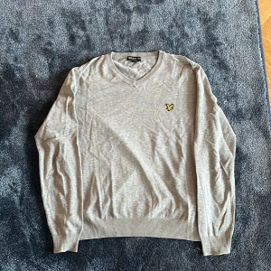 Grå v-ringad tröja från Lyle & Scott - Snygg grå v-ringad tröja från Lyle & Scott med gul broderad logga på bröstet. Tröjan har långa ärmar och ribbade muddar vid hals, ärmslut och nederkant. Perfekt för dig som gillar stilren och klassisk design.