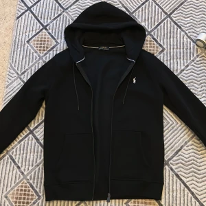 Svart hoodie från Polo Ralph Lauren - Svart hoodie från Polo Ralph Lauren med klassisk vit broderad logga på bröstet. Tröjan har huva med snörning, två fickor fram och dragkedja. Perfekt för chill dagar och har en clean look som funkar till allt.