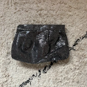Glittrig svart kuvertväska - Säljer en snygg svart kuvertväska med glittrigt tyg och dragkedja upptill. Perfekt storlek för mobilen och det viktigaste. Väskan har en stilren form och glittrande yta som verkligen sticker ut.