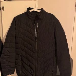 Svart pufferjacka från EA7 Emporio Armani - Svart jacka från EA7 Emporio Armani med hög krage och dragkedja framtill. Jackan har diskret mönstrad quiltning och EA7-logga på dragkedjan. Perfekt för kyliga dagar och ger en clean, sportig look.