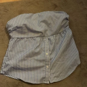 Randig blå tubtopp med knappar - Säljer en blåvit randig tubtopp med knappar framtill. Toppen är axelbandslös och har en bandeau-form med elastisk kant upptill för bättre passform. Perfekt för varma dagar och enkel att styla med jeans eller kjol. 