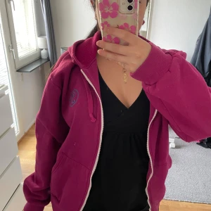 Mörkrosa hoodie  - Säljer en mörkrosa hoodie från Peak Performance med dragkedja och huva. Tröjan har ett blått broderat logomärke på bröstet, snörning i huvan och två stora fickor framtill. Perfekt för dig som gillar en avslappnad och sportig stil.