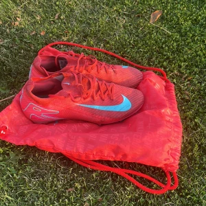 Nike Mercurial röda fotbollsskor - Säljer ett par Nike Mercurial fotbollsskor i röd färg med ljusblå detaljer och snörning. Skorna har en aerodynamisk form, mönstrad ovandel och plös samt dobbar i plast för bra grepp på planen. Snyggt Nike-märke på sidan och levereras med matchande röd påse.
