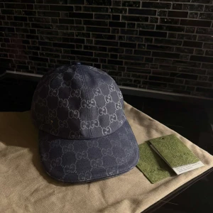 Blå Gucci keps - Snygg blå keps från Gucci med klassiskt GG-monogram över hela kepsen. Justerbar rem baktill i läder och liten guldfärgad GG-detalj på sidan. Kepsen är tillverkad i slitstark bomullscanvas och har en böjd skärm för en sportig look. Perfekt för dig som gillar exklusiva accessoarer.