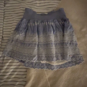 Blå volangkjol med spetsdetaljer - Supersöt blå kortkjol med bred resår i midjan och vackra spetsdetaljer i vitt. Kjolen har volangkant och broderade mönster som ger en romantisk och somrig känsla. Perfekt för dig som gillar en feminin och lekfull stil.