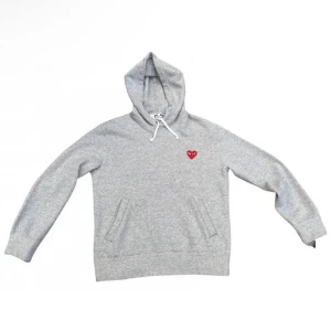Grå hoodie från Comme des Garçons Play - Grå hoodie med huva och snörning från Comme des Garçons Play. Tröjan har ett rött hjärta broderat på bröstet och praktiska fickor framtill. Perfekt för en avslappnad och stilren look.