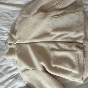 Beige teddyjacka med dragkedja - Aldrig använd!!Mysig beige teddyjacka med hög krage och två stora fickor framtill. Jackan har en hel dragkedja och är fodrad med slätt tyg på insidan. Perfekt för att hålla värmen under kyliga dagar och har en clean, enkel look som funkar till många stilar.