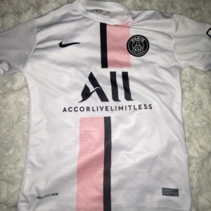PSG Messi 30 vit matchtröja Nike - Snygg PSG fotbollströja från Nike med Messi 30-tryck på ryggen. Tröjan är vit med en rosa och svart vertikal rand på framsidan, PSG-emblem på bröstet och sponsortryck. Tillverkad i lätt och ventilerande material, perfekt för fotbollsplanen eller att bära tröjan på en varm dag. Strl M men kan passar Strl S