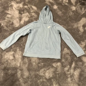 Ljusblå hoodie från Nike i storlek 147-158 - Nike hoodie i ljusblått med klassisk huva och magficka. Tröjan har lång ärm och en enkel, stilren design med Nike-logga på bröstet. Perfekt för chill dagar eller när du vill ha en sportig look.
