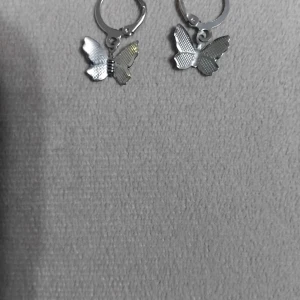 Silvriga örhängen med fjärilar - Ett par snygga örhängen i silverfärg med hängande fjärilar. Fjärilarna har en detaljerad, texturerad yta som ger dem en cool och trendig look. Perfekta för att ge din outfit en extra touch av stil.