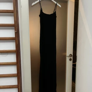 Svart långklänning med smala axelband - En stilren svart långklänning med smala axelband. Klänningen har minimalistisk design och är perfekt för dig som gillar en clean och elegant look. Materialet känns mjukt och följsamt mot kroppen. Aldrig använd. 