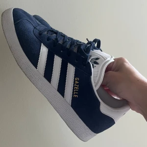 Adidas Gazelle marinblå sneakers - Säljer ett par klassiska Adidas Gazelle sneakers i marinblå mocka med vita detaljer. Skorna har snörning, vit platt sula och vit hälkappa. Använda ca 5 gånger, säljes pga de är för små för mig! 