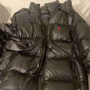 Svart pufferjacka Polo Ralph Lauren M - Svart pufferjacka från Polo Ralph Lauren med glansig finish, röd broderad logga på bröstet och flera dragkedjefickor. Jackan har huva och är vadderad för extra värme. Perfekt för kalla dagar och har en modern, sportig vibe. 