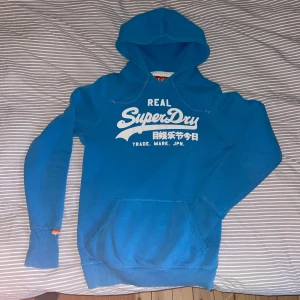 Blå Superdry hoodie med tryck - Säljer en blå hoodie från Superdry med vit logga och japanska tecken på bröstet. Tröjan har huva med snörning, känguruficka och ribbade muddar. Perfekt för dig som gillar streetwear och vill ha en snygg och bekväm tröja till vardags. Färgen är som på 3dje bilden! Skriv vi minsta fundering!🤗
