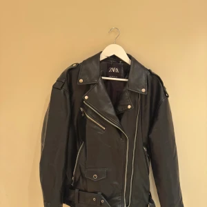 Svart skinnjacka från Zara med bälte - Säljer en svart skinnjacka från Zara i klassisk bikerstil. Jackan har silvriga dragkedjor, axelklaffar, flera fickor och ett bälte i midjan. Snygg och edgy look med detaljer som tryckknappar och bred krage. Perfekt för dig som vill ha en statementjacka.