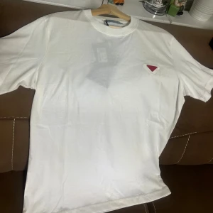 Vit t-shirt från Prada med röd logga - Vit t-shirt från Prada i mjuk bomull med klassisk passform. På bröstet sitter Pradas ikoniska röda trekantslogga i metall. Enkel och clean design med rund hals och korta ärmar. Perfekt för dig som gillar stilrena och exklusiva plagg.