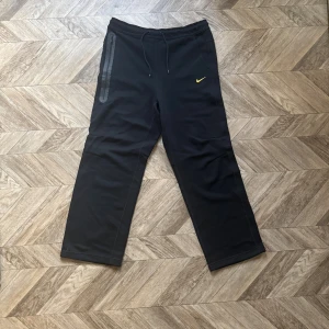 Nocta Nike tech mjukisbyxor - Svarta mjukisbyxor från Nike NOCTA med gul logga och diskreta detaljer. Byxorna har snörning i midjan, raka ben och en dragkedjeficka på sidan. Tillverkade i bomull och polyester för en skön och sportig känsla.