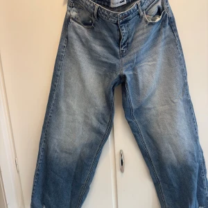 Blå loose jeans från Fashion Nova - Säljer ett par blå jeans från Fashion Nova i loose fit med klassisk femficksdesign och en liten sliten detalj vid fickan. Jeansen är tillverkade i 99% bomull och 1% spandex för lite stretch. Perfekt för dig som gillar en avslappnad och trendig look.