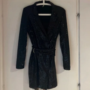 Svart glittrig kortklänning Nelly - Supersnygg svart kortklänning från Nelly med glittriga paljetter över hela klänningen. Långärmad modell med markerad midja och bälte med metallspänne. Perfekt för dig som vill sticka ut med en glamorös look. En liten tråd lös vid sidan.