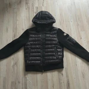 Svart pufferjacka från Moncler - Svart pufferjacka från Moncler med stickade ärmar och vadderad kropp. Jackan har huva, dragkedja framtill och en snygg ficka med dragkedja på ärmen. Moncler-logga på vänster ärm. Perfekt för dig som vill ha en stilren och sportig look.