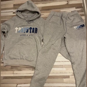 Trapstar hoodie och byxor set  - Säljer en grå hoodie från trapstar med blått och vitt tryck framtill. Tröjan har huva med snörning och en stor magficka. Perfekt för chill dagar och matchar snyggt med mjukisbyxor. Mjuk och skön bomullskänsla.