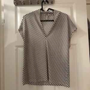 Randig blus med v-ringning från H&M - Snygg blus från H&M i vitt med svarta diagonala ränder. Modellen har korta ärmar, v-ringning och en lös passform. Perfekt att styla med jeans eller kjol för en trendig look. Materialet känns mjukt och lätt, vilket gör den skön att bära hela dagen.