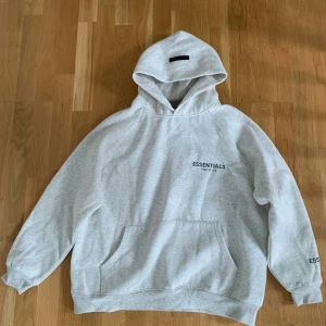 Grå Essentials Fear of God hoodie - Säljer en ljusgrå hoodie från Essentials Fear of God. Tröjan har huva, känguruficka och tryck på bröstet samt ärmen. Oversized passform och mjukt material som känns riktigt skönt. Perfekt för dig som gillar streetwear och enkel stil.