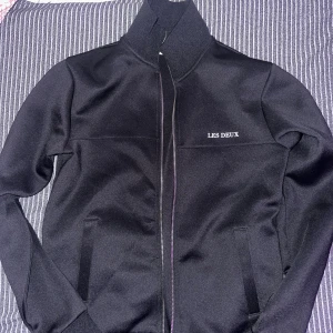 Svart jacka från Les Deux XS - Snygg svart jacka från Les Deux i storlek XS. Jackan har hög krage, dragkedja framtill och två fickor. Enkel och stilren design med diskret logga på bröstet. Perfekt för dig som gillar en clean och sportig look. VARAN ÄR VÄLDIGT STOR I STORLEK. JAG ÄR 187 och den passar mig