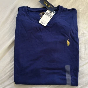 Blå t-shirt från Polo Ralph Lauren - Snygg marinblå t-shirt från Polo Ralph Lauren med klassisk gul broderad logga på bröstet. T-shirten är i bomull och har rund halsringning och korta ärmar. Perfekt för en clean och stilren look.