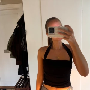 Svart halterneck tubtopp - svart halterneck tubtopp med breda band som knyts i nacken. Toppen är croppad och har en tight passform med lätt rynkade detaljer längs sidorna. Perfekt att styla med jeans eller kjol för en trendig look.