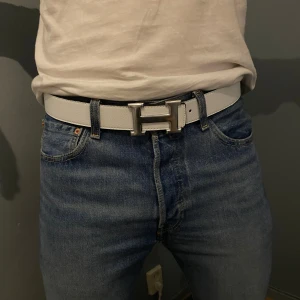 Vändbart bälte med H-spänne - Säljer ett vändbart H-bälte i med ikoniskt H-spänne i silver. Ena sidan är vit och andra svart, så du kan matcha med olika outfits. Bältet har stilren design och passar perfekt till jeans eller kostymbyxor.