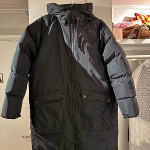 Peak Performance W Stella Coat - kanonskick! - Peak Performance W Stella Coat i svart, lång modell med huva och stora fickor framtill. Jackan har dragkedja hela vägen, snörning i huvan och smarta ventilationsdragkedjor i sidorna. Perfekt för kalla dagar. Sista bilden är lånad från Peak för att visa från sidan. Ord.pris 7000kr