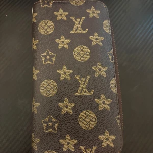 plånbok med brunt skinn - Säljer en brun plånbok från med klassiskt monogrammönster i guld. Plånboken är tillverkad i skinn och har dragkedja runtom. Perfekt för dig som vill ha en snygg och lyxig accessoar i väskan.