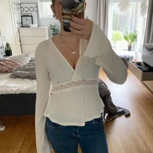 Vit omlottopp med spetsdetalj - Supersöt vit omlottopp med lång ärm och v-ringning. Toppen har en bred spetsdetalj i midjan med blommigt mönster och lätt utsvängd nederdel. Tunt, flowy material som ger en romantisk vibe och extra långa ärmar för en cool look. Har Andvänt 1 gång.