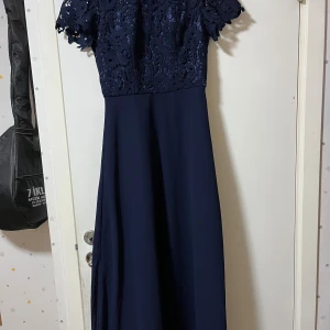 Marinblå långklänning med spets - Elegant marinblå långklänning med kortärmad överdel i vacker spets och lång, slät kjol. Klänningen har rund halsringning och en feminin siluett som ger en klassisk look. Perfekt för dig som vill sticka ut med stilren och tidlös design.