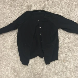 Svart kofta från Next med knappar - En klassisk svart kofta från Next med rund halsringning och knäppning framtill. Långärmad och i mjukt material som passar perfekt till lager-på-lager. Stilren och enkel design som funkar till det mesta.