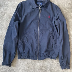 Marinblå vindjacka Polo Ralph Lauren XS - Snygg marinblå vindjacka från Polo Ralph Lauren i 100% bomull. Klassisk design med krage, dragkedja framtill och två sidofickor. Jackan har elastisk nederkant och är prydd med röd Polo-logga på bröstet. Perfekt för dig som gillar stilrena och tidlösa plagg.