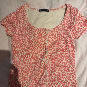 Leopardmönstrad topp från Brandy Melville - Säljer en kortärmad topp från Brandy Melville med rosa leopardmönster på ljus bakgrund. Toppen har rund halsringning och knappar framtill. Perfekt för dig som gillar färgglada och trendiga prints.
