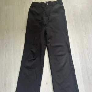 Svarta kostymbyxor med raka ben - Svarta kostymbyxor med klassisk rak passform och pressveck. Byxorna har hällor för bälte, diskreta fickor och stängs med knapp och dragkedja. Perfekta för en clean och stilren look.